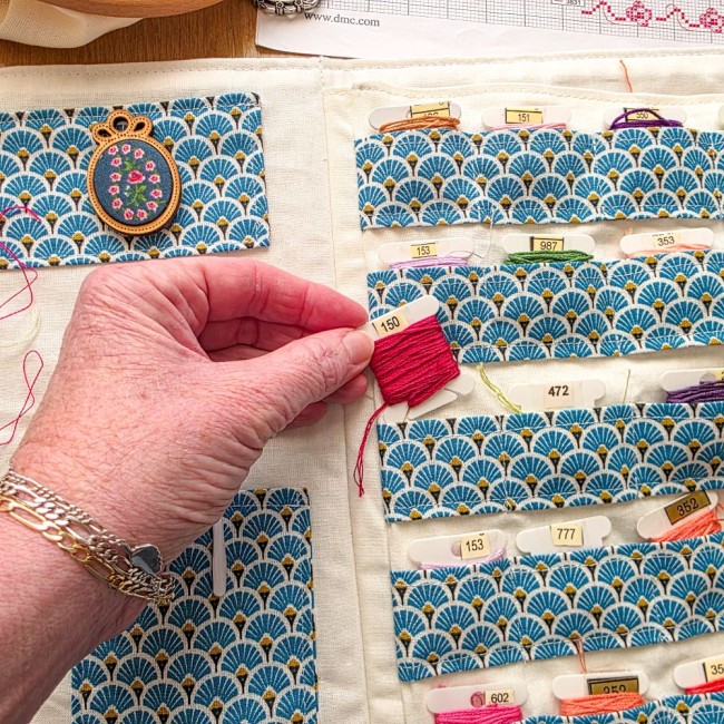 trousse-brodeuse-rangement-cartonnettes-fils-mains-et-merveilles-angelique-la-fee-en-fil Main saisissant une cartonnette de fil rose dans les rangements de la trousse de brodeuse Mains et Merveilles aux motifs éventails bleus et dorés, création artisanale Angélique La Fée En Fil.