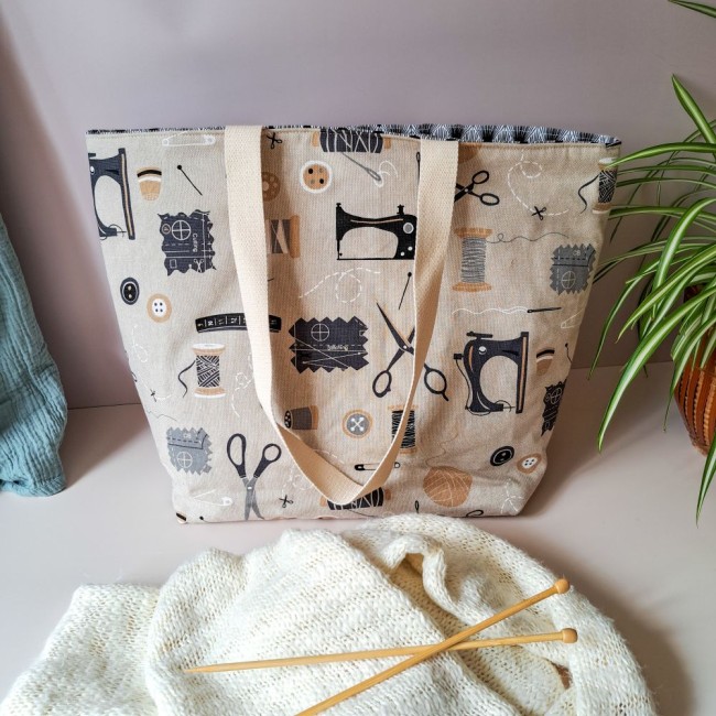 sac-ouvrage-tricot-angelique-la-fee-en-fil Posé sur la table près d'une plante, un sac à ouvrage Louise pour tricot, de couleur beige avec des motifs de machine à coudre. Devant un tricot et des aiguilles à tricoter. Création Angélique La Fée En Fil