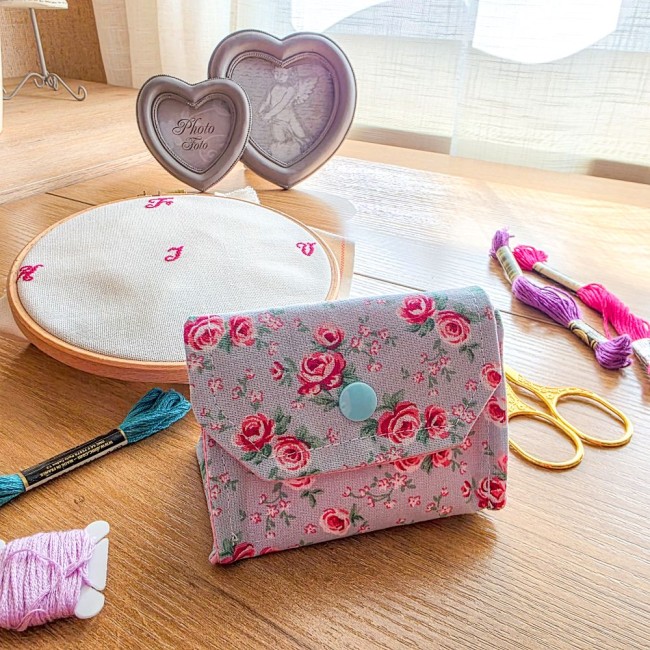 Vue de face d'une mini poubelle à fils pliable fermée pour broderie et point de croix, modèle Bianca en tissu gris clair aux motifs bucoliques de roses rouges avec rabat et bouton-pression bleu clair, posée sur une table en bois à côté d'un tambour à brod