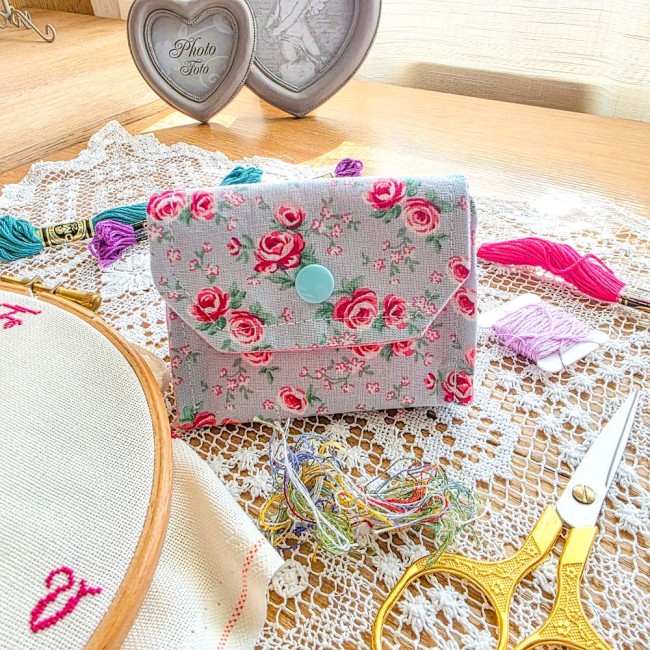 Mini poubelle à fils pliable en tissu bleu ciel imprimé de roses rouges et de feuillages avec bouton-pression bleu turquoise. Posée sur un napperon en dentelle avec accessoires de broderie, création artisanale Angélique La Fée En Fil.