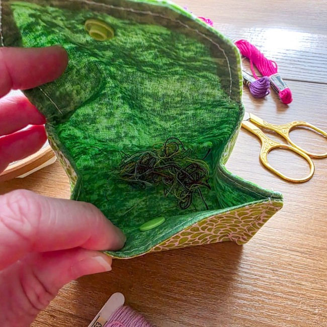Gros plan de l'intérieur d'une poubelle à fils de table modèle Verveine montrant la doublure verte remplie de chutes de fils de broderie, tenue en main avec ciseaux dorés et échevettes en arrière-plan, création artisanale Angélique La Fée En Fil.