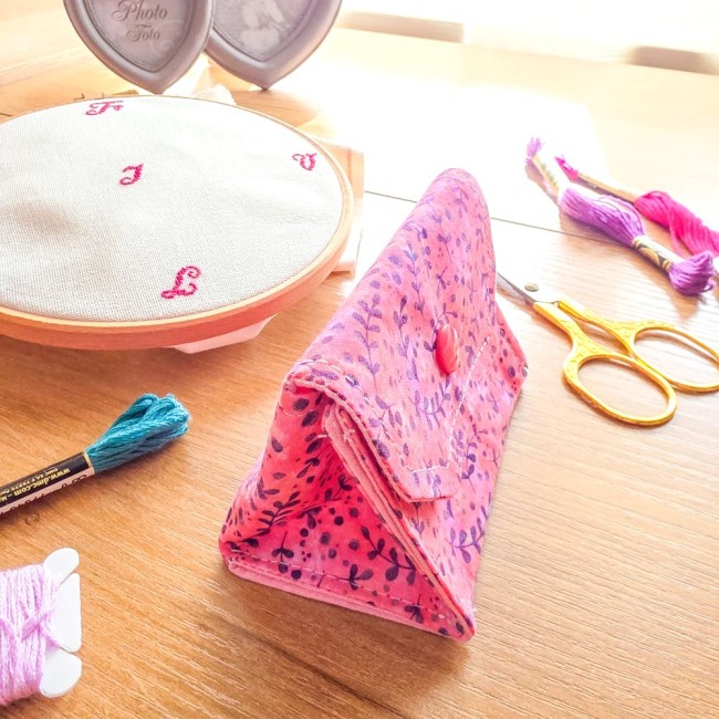 Vue de trois-quarts d'une mini poubelle à fils pliable fermée pour broderie et point de croix, modèle Sandy Rose en tissu rose aux motifs de feuillage violet avec rabat et bouton-pression rose, posée sur une table en bois à côté d'un tambour à broder, de 