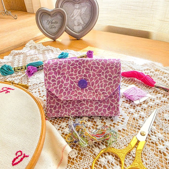 Mini poubelle à fils pliable en tissu mauve imprimé de petites gouttes blanches avec bouton-pression violet, posée sur un napperon en dentelle avec accessoires de broderie, création artisanale Angélique La Fée En Fil.