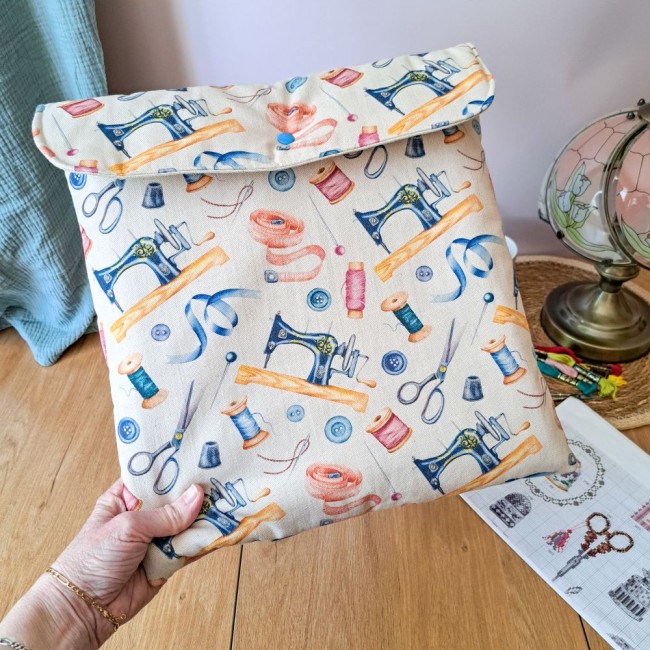 Main tenant une grande pochette de rangement pour broderie molletonnée, fermée par un bouton pression bleu. Le tissu crème présente des illustrations aquarelle de machines à coudre anciennes, rubans et bobines de fil. En arrière-plan, une lampe et une gri