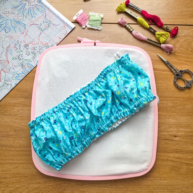 grime-guard-sur-cadre-nurge-rose-bleu-turquoise-fleuri-angelique-la-fee-en-fil Un couvre-cadre (grime guard) élastiqué en coton bleu turquoise imprimé de petites fleurs blanches, bleues et jaunes, posé en diagonale sur une toile Aïda blanche tendue dans un cadre carré rose de marque Nurge. L'étiquette de la marque est visible. En ar