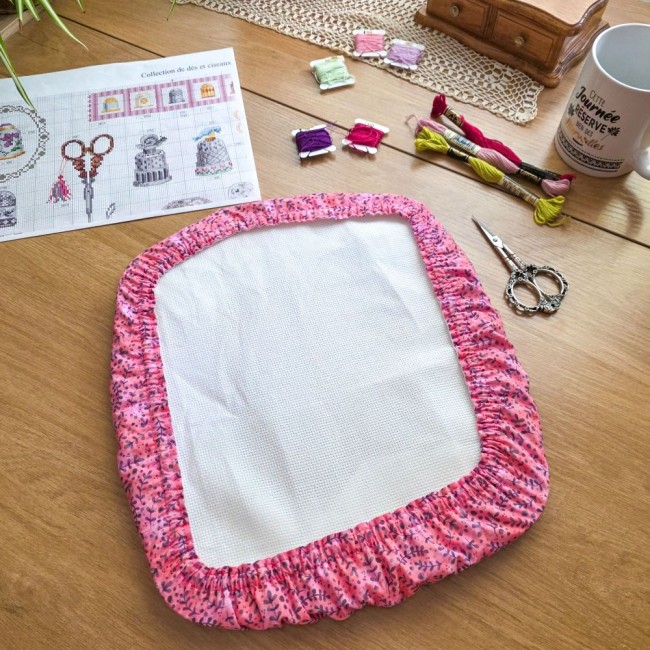 grime-guard-sous-bois-rose-cadre-nurge-broderie-angelique-la-fee-en-fil Un couvre-cadre (grime guard) modèle Sous-bois Rosé en coton vieux rose à motifs de branches violettes, épousant parfaitement un cadre à broder carré de marque Nurge avec sa toile Aïda blanche. La mise en scène sur une table en bois comprend un diagramme