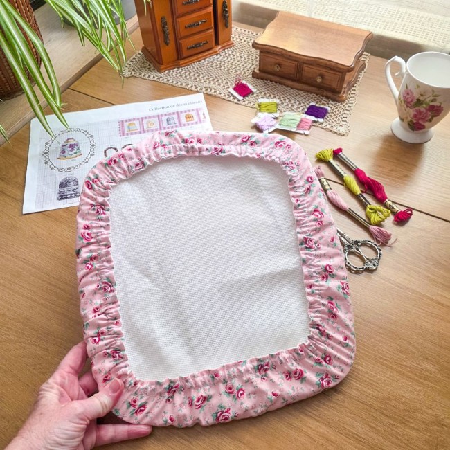 grime-guard-cadre-rectangulaire-main-point-de-croix-romane-angelique-la-fee-en-fil Gros plan des mains tenant un grime guard housse protège-toile modèle Romane installé sur un grand cadre à broder rectangulaire, en tissu rose aux motifs bucoliques de fleurs et roses, avec ciseaux vintage, échevettes et grille de point de croix en arrièr