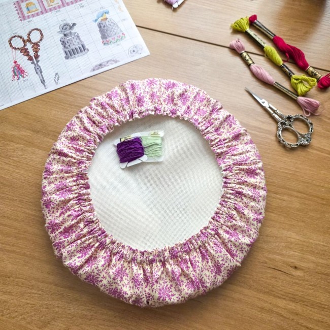 Vue d'ensemble d'un grime guard housse protège-toile modèle Samba en tissu coton à motifs floraux rose et violet, installé sur un tambour à broder rond avec échevettes, ciseaux vintage et grille de broderie en arrière-plan, création artisanale Angélique L