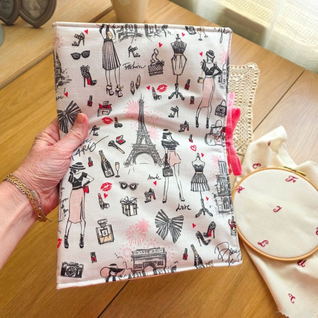 Main tenant la trousse de brodeuse Nina fermée, montrant sa couverture en tissu imprimé d'illustrations parisiennes et de mode avec la Tour Eiffel et un ruban rose, création artisanale Angélique La Fée En Fil.