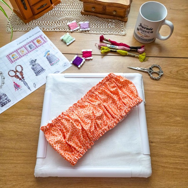 grime-guard-cadre-q-snap-carre-point-de-croix-mandarine-orange-angelique-la-fee-en-fil Vue d'ensemble d'un grime guard housse protège-toile modèle Mandarine en tissu coton orange imprimé de motifs gouttelettes blanches, ajusté sur un cadre carré de type Q-Snap, avec grille de broderie, ciseaux, échevettes de fils et mug sur une table en boi
