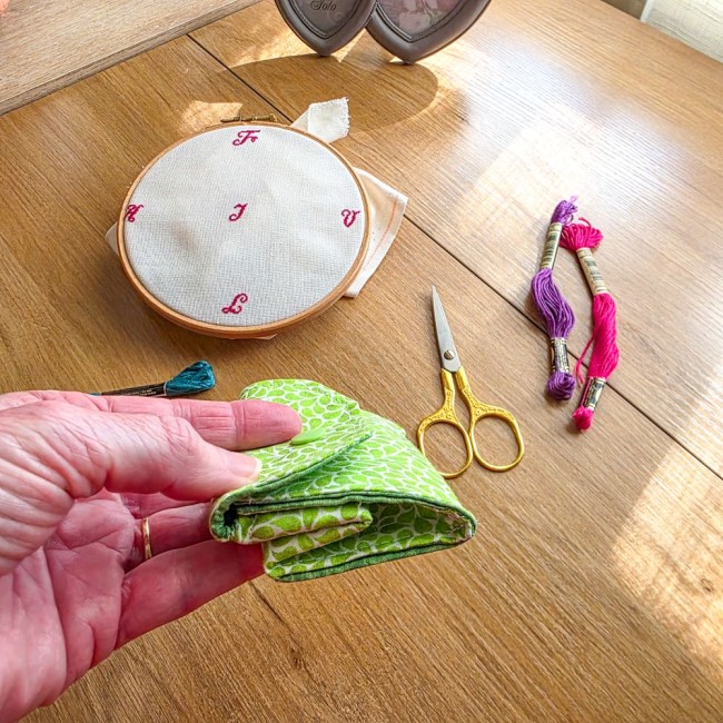 Gros plan d'une main tenant une poubelle à fils de table repliée à plat modèle Verveine avec pression verte, en tissu vert anis aux motifs de feuillage blanc, avec ciseaux dorés, tambour à broder et échevettes de fils en arrière-plan, création artisanale 