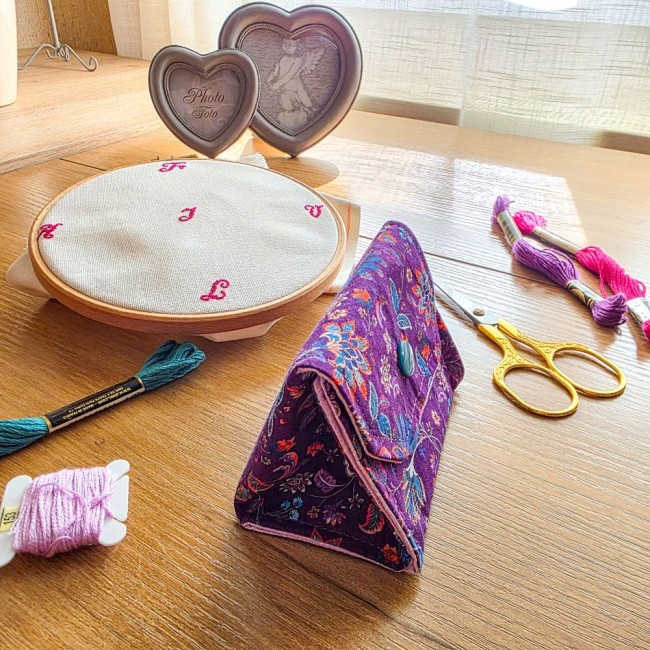 Vue de profil de la mini poubelle à fils nomade et pliante modèle Cachemire en tissu violet aux motifs floraux et cachemire, montrant sa forme triangulaire sur le côté et son rabat fermé, avec tambour à broder, ciseaux dorés et échevettes, création artisa