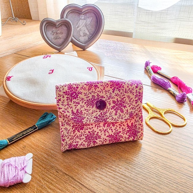 Vue de face d'une mini poubelle à fils de table pliante fermée modèle Samba en tissu coton à motifs floraux rose et violet avec bouton-pression violet assorti, posée sur une table en bois avec accessoires de broderie, création artisanale Angélique La Fée 