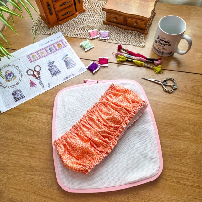 grime-guard-cadre-nurge-broderie-mandarine-orange-angelique-la-fee-en-fil Vue d'ensemble d'un grime guard housse protège-toile modèle Mandarine en tissu coton orange imprimé de motifs gouttelettes blanches, ajusté sur un cadre rectangulaire rose de type Nurge, avec grille de broderie, ciseaux, échevettes de fils et mug sur une