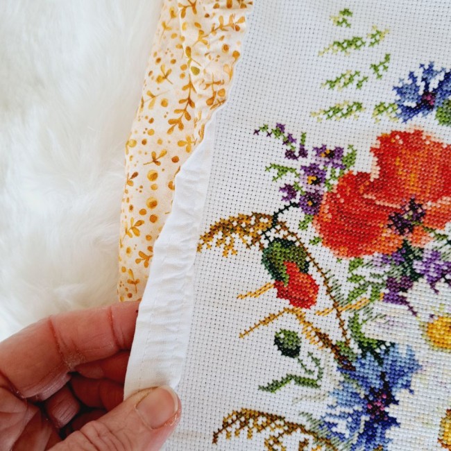 grime-guard-sous-bois-dore-detail-bordure-elastique-broderie-angelique-la-fee-en-fil Gros plan d'une main soulevant le bord élastique blanc d'un grime guard modèle Sous-Bois Doré en tissu beige à motifs de feuillage ocre sur une toile brodée au point de croix par Angélique La Fée en Fil