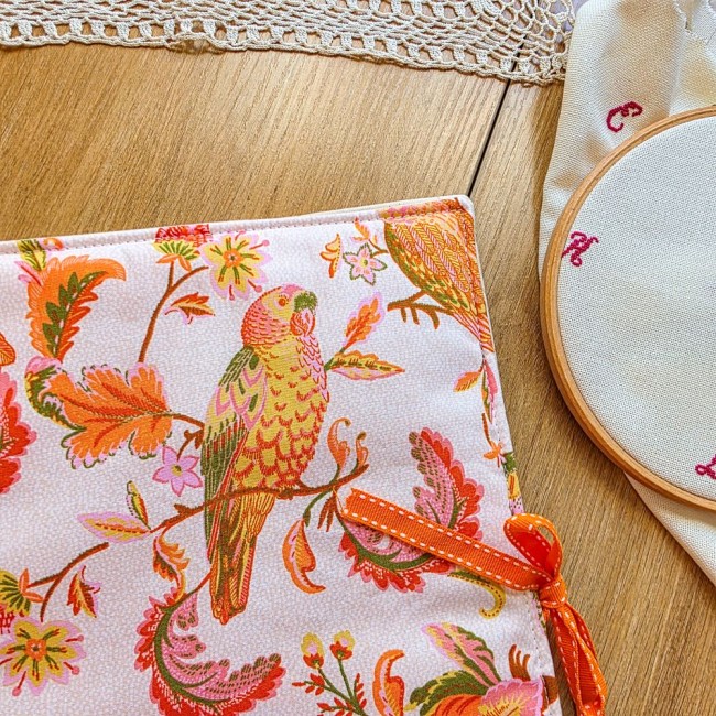 Gros plan sur le tissu de la trousse de broderie Savannah montrant un perruche ou perroquet perché sur une branche fleurie et le lien en ruban orange, création artisanale Angélique La Fée En Fil.