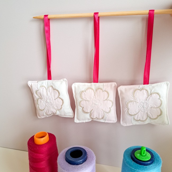 bijou-ciseaux-brodeuse-coussinet-fleur-blanche-ruban-rose-fils-bobines-atelier-angelique-la-fee-en-fil Trois bijoux de ciseaux pour brodeuse en forme de coussinets blancs brodés d’une fleur, suspendus par des rubans roses, avec des bobines de fil colorées en arrière-plan, création artisanale Angélique La Fée En Fil.