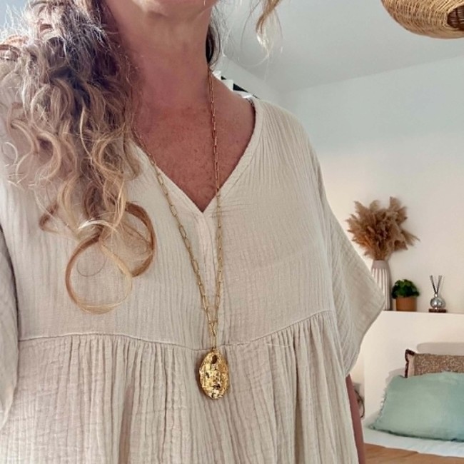 Collier long Mélusine doré porté par une femme Sautoir Mélusine doré porté avec pendentif ovale mrtelé sur robe écrue