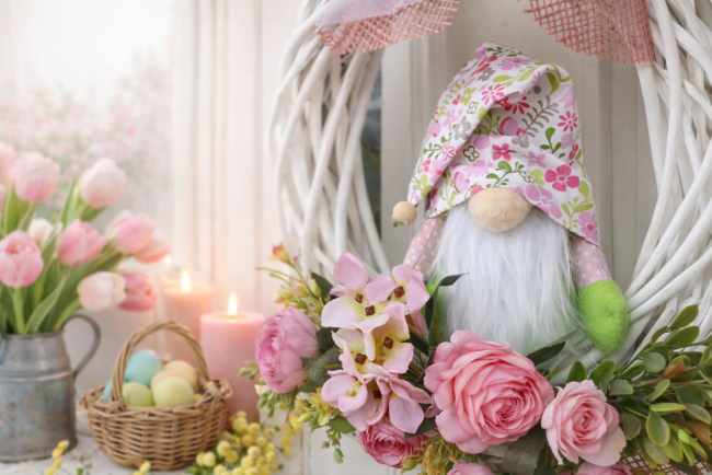 Gros plan sur gnome artisanal au chapeau fleuri rose dans couronne florale blanche, décorée de roses et feuillage vert, ambiance douce de printemps.