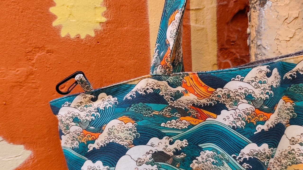 un beau sac suspendu dans la rue devant un mur terra cotta avec un soleil jaune. On peut voir une dragonne avec un petit mousqueton noir. Les motifs sont la mer avec des vagues scélérates et le mont fuji inspiration hokusai en bleu et vert canard orange et écru