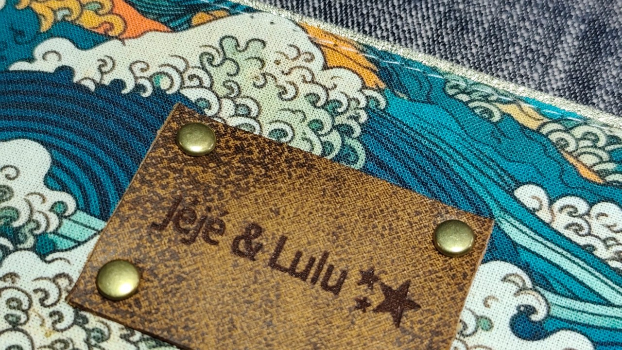 Une étiquette rectangulaire en simili cuir gravée avec marqué Jéjé & Lulu accompagné de petites étoiles. Elle est posée avec des rivets dorés clair sur un beau tissu à motif japonais la vague de Kanagawa aux tons bleu sarcelle, orange, blanc. On aperçoit un passepoil doré et du chambray
