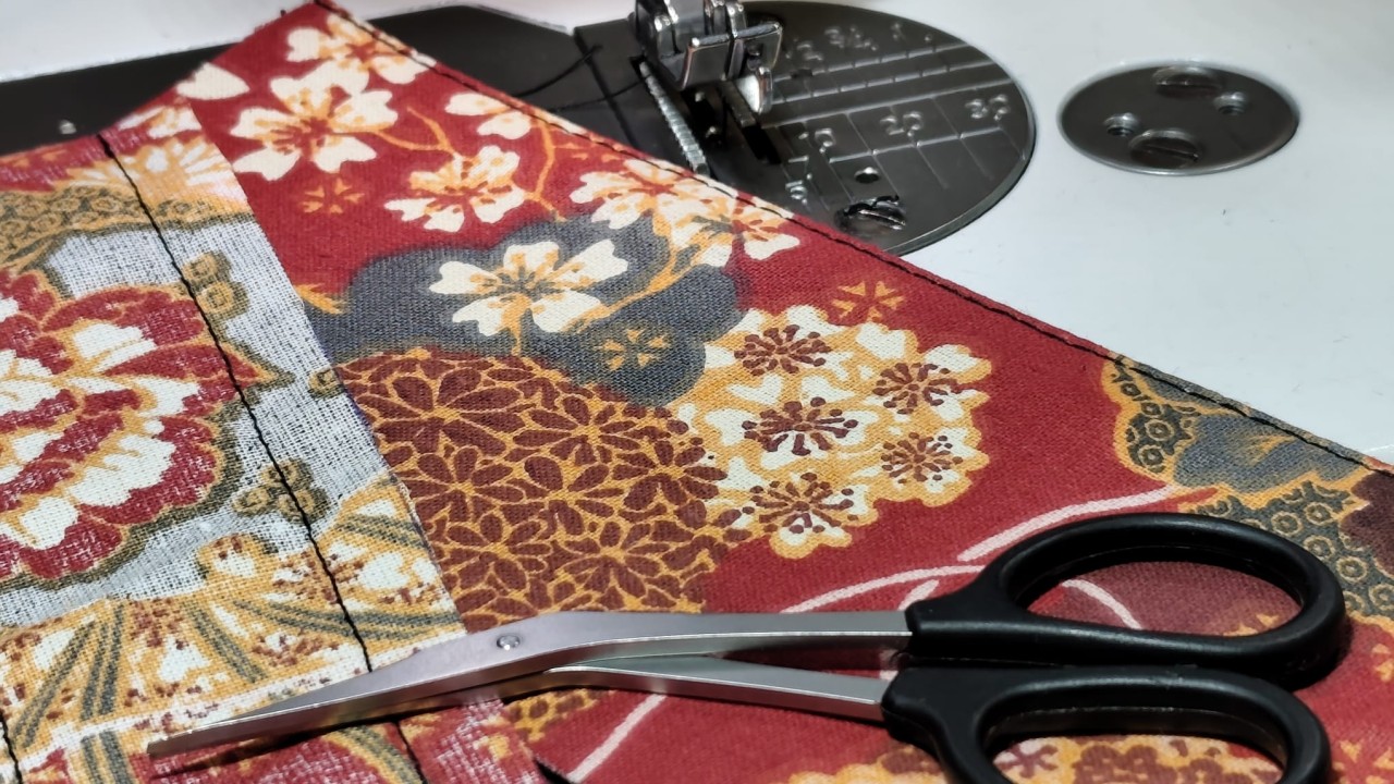 une paire de petits ciseaux de couture posées sur un beau tissu japonais à motif fleuri et coloré de rouge, jaune vert et blanc. Le tout est posé sur une machine à coudre. On peut voir aussi une couture droite avec du fil noir.
