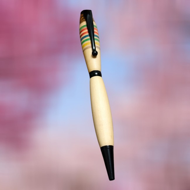 un stylo bille au milieu de l'image en buis avec un motif rayures multicolores comme sur une planche de skate et une incrustations sur le haut avec du bois marron foncé qui a un veinage bien prononcé. Il est monté sur un kit métal noir. Le fond de l'image