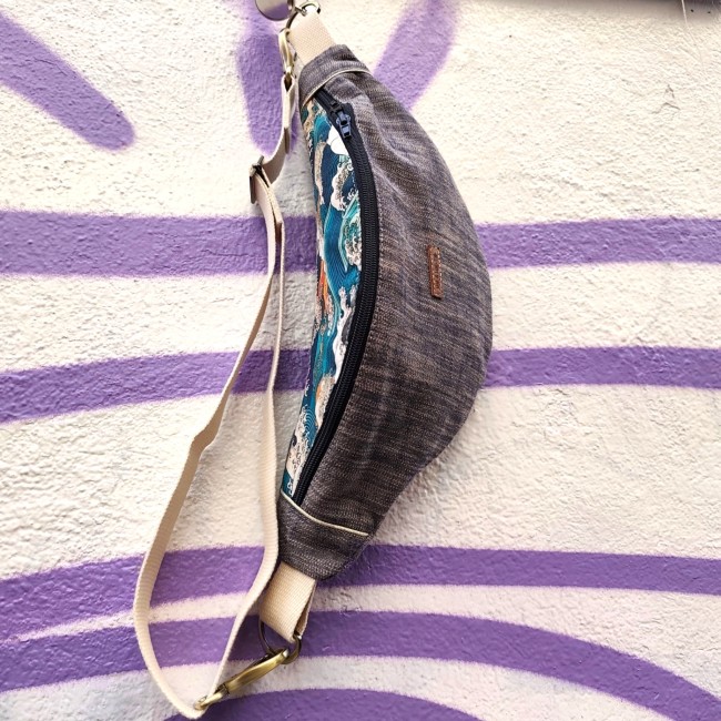 sac banane en tissu bi colore et bi matière avec une sangle coton écru et des boucles métal étain. Le tissu à motif est de couleur bleu sarcelle, orange et écru. Suspendu à un mur blanc avec un graffiti violet.