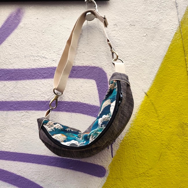 sac banane en tissu bi colore et bi matière avec une sangle coton écru et des boucles métal étain. Le tissu à motif est de couleur bleu sarcelle, orange et écru. Suspendu à un mur blanc avec un graffiti violet.