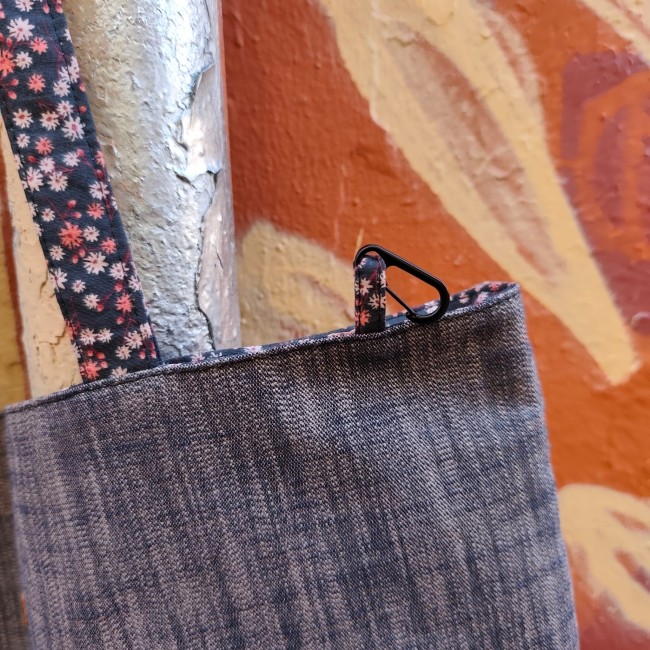 Accessoires textiles de qualité fabriqués à Marseille - Artisanat local - Jéjé & Lulu zoom sur un haut de sac cabas en chambray et tissu à petites fleurs roses avec sa dragonne munie d'un petit mousqueton en métal noir. Devant un tuyau gris métallisé et un mur ocre peint de feuilles.