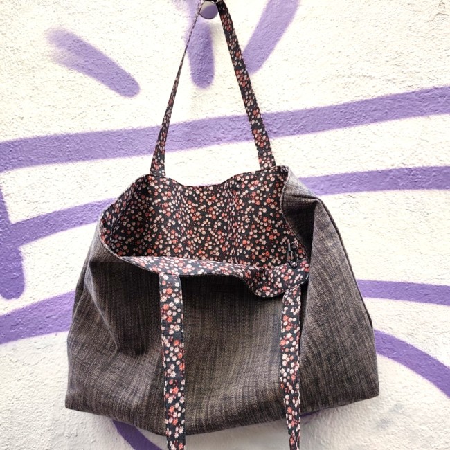 Accessoires textiles de qualité fabriqués à Marseille - Artisanat local - Jéjé & Lulu un sac en tissu réversible ouvert on peut voir du chambray pour l'extérieur et du coton fleuri pour l' intérieur et les anses. Il est suspendu devant un mur blanc avec des graffitis violet.
