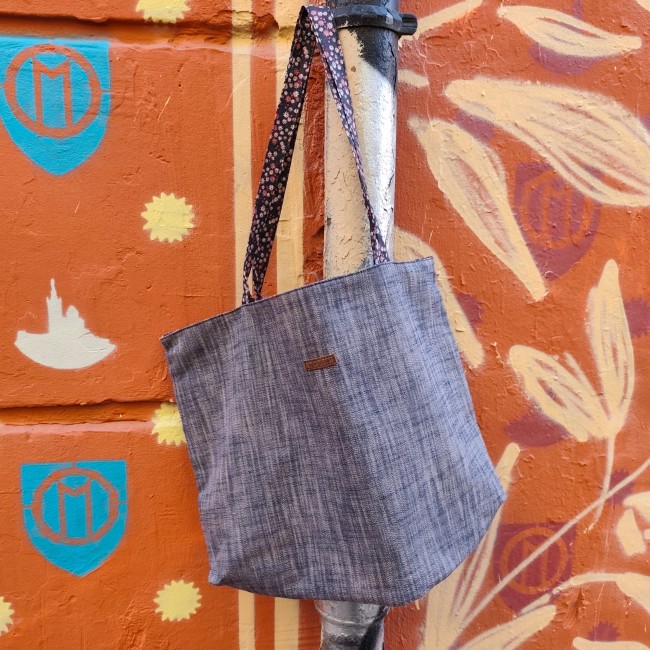 Accessoires textiles de qualité fabriqués à Marseille - Artisanat local - Jéjé & Lulu Un beau sac en chambray réversibl avec des anses fleuries suspendu à un tuyau style zèbre noir et blanc devant un mur ocre avec un logo om bleu turquoise, un dessin de notre dame de la garde et des soleils . Photo prise au cours julien, quartier street ar