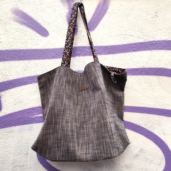 Accessoires textiles de qualité fabriqués à Marseille - Artisanat local - Jéjé & Lulu vue de devant d'un sac en chambray avec des anses fleuries et une étiquette en simili cuir marron gravée jéjé & lulu suspendu à un mur blanc avec un tag violet
