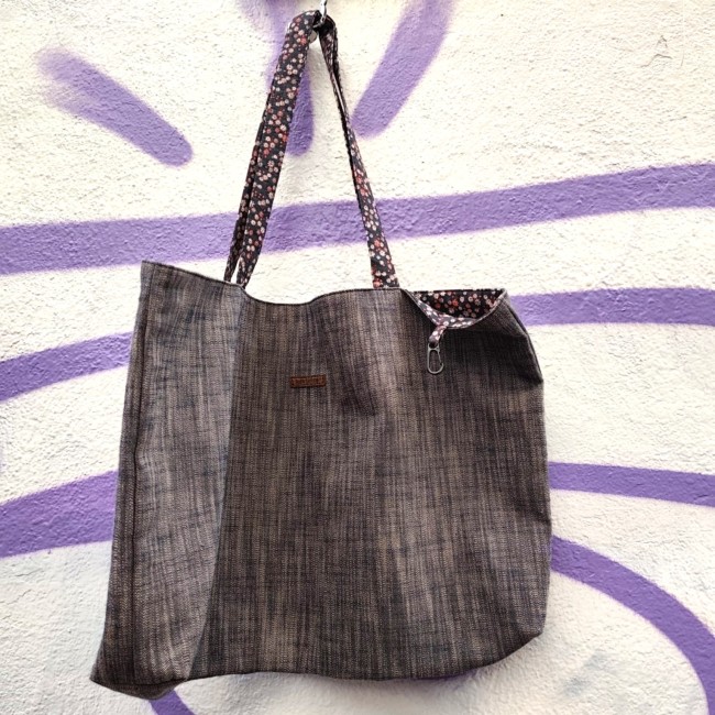 Accessoires textiles de qualité fabriqués à Marseille - Artisanat local - Jéjé & Lulu vue de devant d'un sac en chambray avec des anses fleuries et une étiquette en simili cuir marron gravée jéjé & lulu suspendu à un mur blanc avec un tag violet