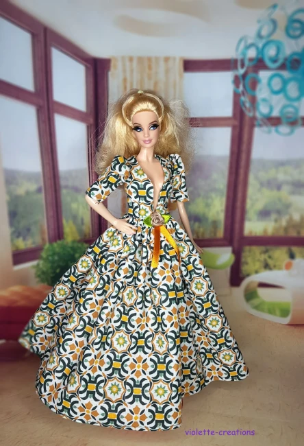 Robe Galla Mosaïque Vert & Ocre Poupée | Violette-Creations Robe de galla longue pour poupée type Barbie en coton, motif mosaïque géométrique vert émeraude et ocre sur fond écru, décolleté V, manches bouffantes, blason doré et ruban de velours ocre à la taille, création artisanale par Violette-Creations.
