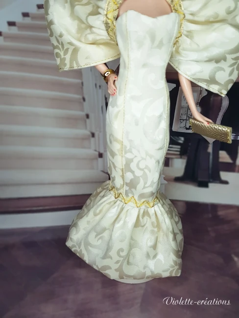 Poupée Barbie de collection en robe de soirée sirène dorée et argentée, posant dans un salon de luxe feutré avec chandeliers.