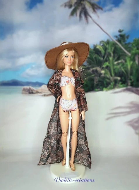 Poupée type Barbie portant un maillot de bain deux pièces pastel avec kimono fleuri transparent, chapeau de soleil et sandales sur un décor de plage grecque.