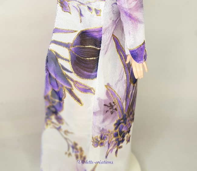 Poupée mannequin type Barbie portant un maillot de bain fleuri lavande avec kimono de plage et chapeau assorti dans un décor tropical.