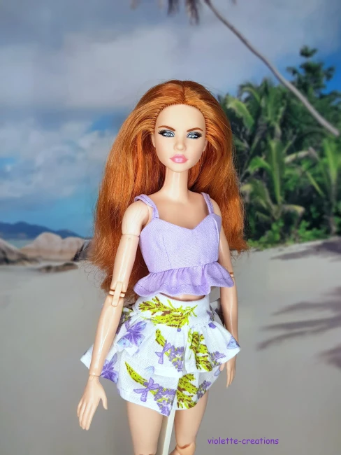 Vêtement pour poupée Barbie composé d'un haut court lilas et d'une jupe blanche à motifs fleurs violettes et feuilles vertes sur fond de plage.