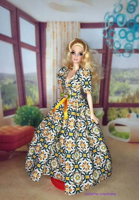 Robe de galla longue pour poupée type Barbie en coton, motif mosaïque géométrique vert émeraude et ocre sur fond écru, décolleté V, manches bouffantes, blason doré et ruban de velours ocre à la taille, création artisanale par Violette-Creations.