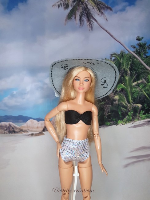 Maillot de bain 2 pièces pour poupée mannequin type Barbie avec haut noir, bas argenté et chapeau en feutrine sur décor de plage.