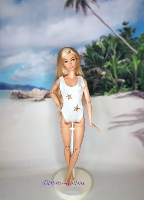 Maillot de bain 1 pièce blanc et étoiles dorées pour poupée mannequin type barbie (2) Maillot de bain 1 pièce en lycra blanc avec deux étoiles dorées pailletées sur une poupée mannequin type Barbie blonde, devant un fond de plage tropicale.