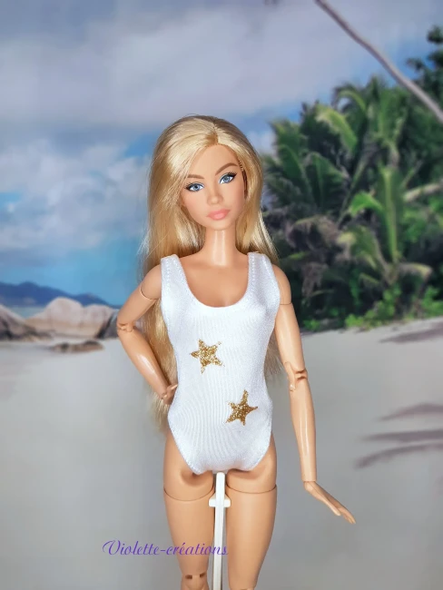 Maillot de bain 1 pièce blanc et étoiles dorées pour poupée mannequin type barbie (3) Maillot de bain 1 pièce en lycra blanc avec deux étoiles dorées pailletées sur une poupée mannequin type Barbie blonde, devant un fond de plage tropicale.