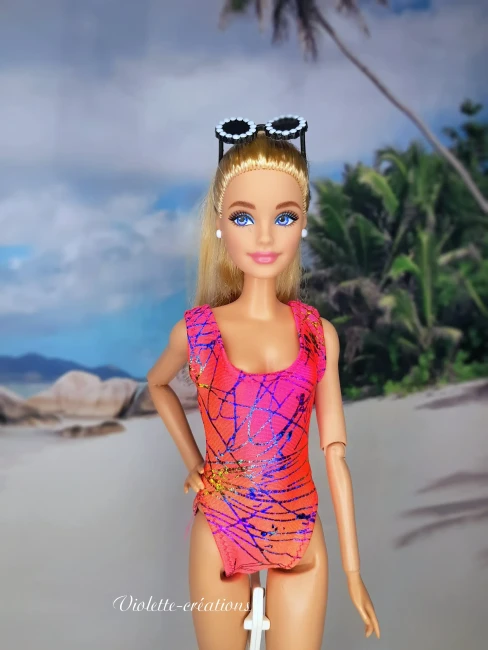 Maillot de bain 1 pièce graphique rose et orange pour poupée mannequin type barbie (3) Maillot de bain 1 pièce en lycra rose et orange à motifs graphiques sur une poupée mannequin blonde avec lunettes de soleil, devant un fond de plage tropicale.