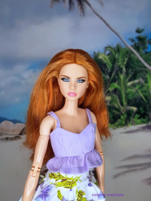Vêtement pour poupée Barbie composé d'un haut court lilas et d'une jupe blanche à motifs fleurs violettes et feuilles vertes sur fond de plage.