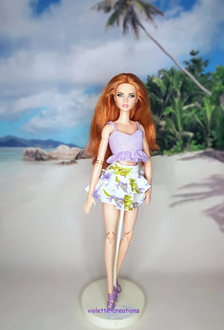 Vêtement pour poupée Barbie composé d'un haut court lilas et d'une jupe blanche à motifs fleurs violettes et feuilles vertes sur fond de plage.