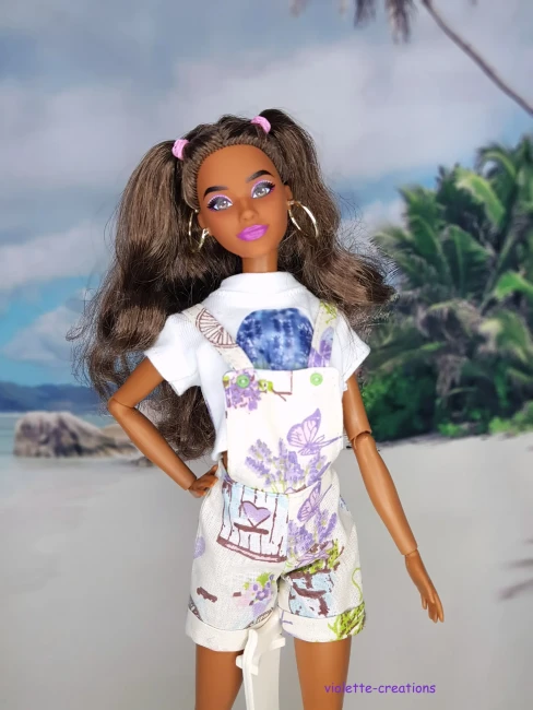 Poupée Barbie portant une salopette short style Shabby Chic avec motifs lavande et papillons, accompagnée d'un crop-top blanc illustré, création artisanale Violette-Créations sur un fond de plage tropicale.