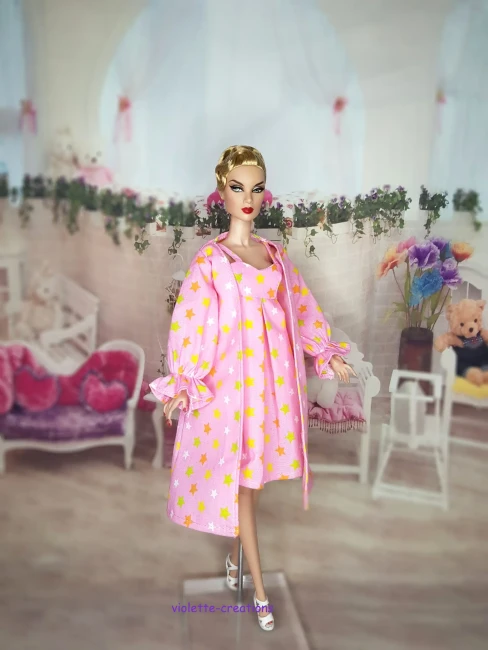 Ensemble Nuit Poupée Barbie : Nuisette Rose et Peignoir Étoiles Poupée Barbie portant un ensemble de nuit rose avec motif étoiles jaunes et blanches, incluant une robe de chambre à volants.