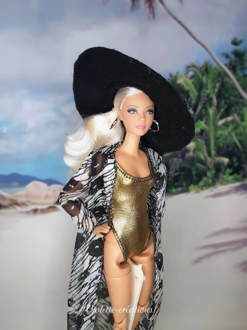 Poupée mannequin blonde portant un maillot de bain une pièce en lycra doré, un long kimono de plage en crêpe à motifs noirs et blancs avec liseré doré, et un grand accessoire en feutrine noire, posant devant un décor de plage tropicale. 