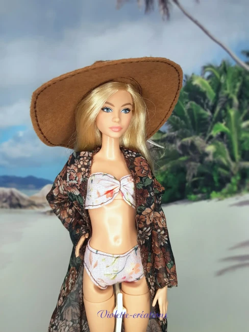 Poupée type Barbie portant un maillot de bain deux pièces pastel avec kimono fleuri transparent, chapeau de soleil et sandales sur un décor de plage grecque.